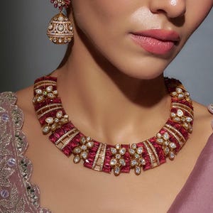 Op de afbeelding: Een gouden ketting met een rood en wit stenen ontwerp. De ketting is gemaakt van goud en heeft een geometrisch patroon. De stenen zijn ingelegd in een bloemmotief.