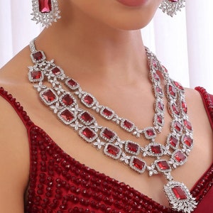 Puede incluir: Un collar de plata de tres hileras con detalles de piedras preciosas rojas. El collar presenta un colgante con una piedra preciosa roja grande y piedras preciosas más pequeñas que la rodean. El collar lo lleva una persona con una blusa roja de cuentas.