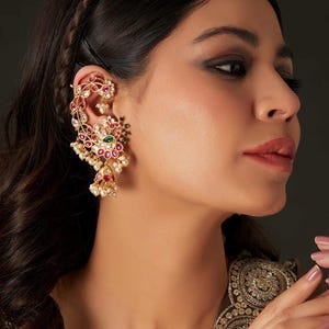 May include: An ornate gold ear cuff featuring a floral and leaf design, adorned with red, green, and white gemstones, and small white pearls. The jewelry piece is worn on the ear, showcasing its intricate details and craftsmanship.
