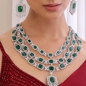 Peut inclure: Un ensemble de collier à trois rangs et boucles d'oreilles assorties. Les bijoux présentent de grandes pierres vertes émeraude de coupe carrée, entourées de petites pierres rondes et claires. Le collier a un pendentif avec une grande pierre verte émeraude.