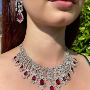 Può includere: Una collana d'argento con un design a forma di lacrima presenta pietre preziose rosse e strass trasparenti. La collana è abbinata a orecchini coordinati.