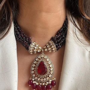 May include: A gold necklace with a large red gemstone pendant surrounded by smaller white gemstones. The necklace is made of multiple strands of dark red beads.