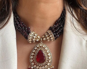 May include: A gold necklace with a large red gemstone pendant surrounded by smaller white gemstones. The necklace is made of multiple strands of dark red beads.