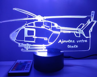 May include: A clear acrylic helicopter-shaped LED lamp with blue illumination, mounted on a round black base. The design features the French text "Ajoutez votre texte" etched into the acrylic. A small rectangular remote control is included.