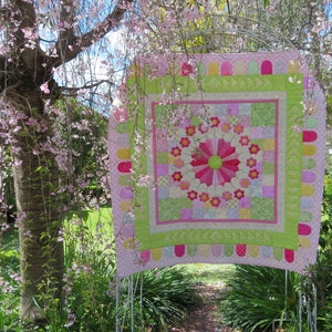 May include: A pink and green patchwork quilt with a large flower design in the center. The quilt is hanging from a tree branch in a garden setting.