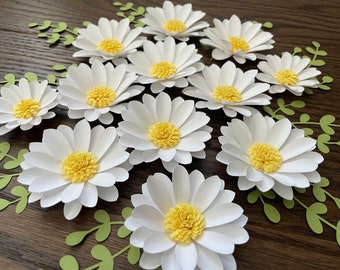 May include: A collection of 10 white paper daisies with yellow centers. The daisies are arranged in a circle on a brown wooden surface.
