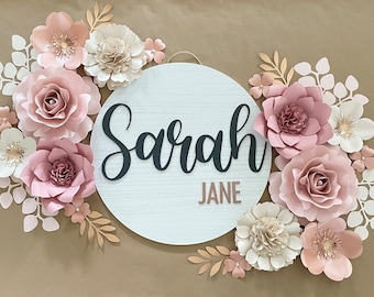 May include: A white round wooden sign with the name "Sarah Jane" written in black script. The sign is decorated with pink and white paper flowers.
