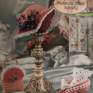 Puede incluir: Un sombrero, un bolso y un zapato de ganchillo en rosa y blanco con un soporte decorativo. La imagen es de Annie's Miniature Shoe Society, una colección llamada "Stepping Through Time: A Lacy Afternoon".