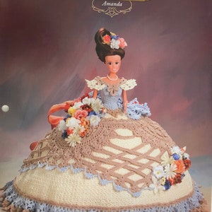 May include: A crochet doll wearing a white and brown dress with a blue lace trim. The doll has brown hair and is wearing a flower crown. The doll is holding a bouquet of flowers. The image is from Annie's Glorious Gowns Old South Collection, Amanda.