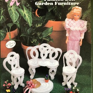 May include: A white plastic canvas dollhouse furniture set with a table and four chairs. The set is designed to be made by hand using plastic canvas and yarn. The furniture is white and has a delicate, ornate design. The set is shown on a green background with a doll and flowers.