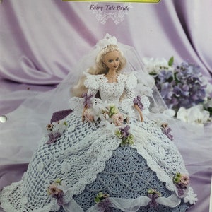 May include: A doll wearing a white crocheted wedding gown with a veil. The dress features a detailed openwork design, adorned with small floral accents in shades of pink and purple. The text "Annie's Glorious Gowns" and "Flower Garden Collection" are visible.