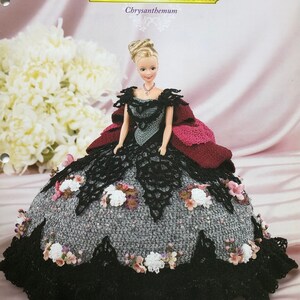 May include: A doll in a handmade gown from the "Flower Garden Collection." The dress has a black lace bodice and a grey, textured skirt with white and pink floral accents. The text "Annie's Glorious Gowns" is visible.