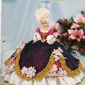 Könnte beinhalten: Eine Puppe in einem gehäkelten Kleid aus der "Flower Garden Collection". Das Kleid hat einen dunkelblauen Rock mit goldenem Besatz, ein rotes Oberteil und weiße Spitzenärmel. Die Puppe hat eine große blonde Perücke und ist mit rosa Blumen verziert.