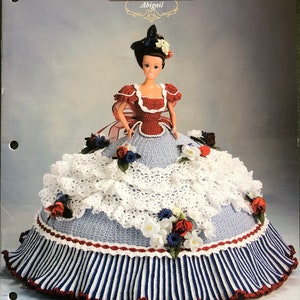 May include: A crochet doll dress pattern for a doll wearing a red and white dress with a blue and white striped skirt. The pattern is titled "Annie's Glorious Gowns Old South Collection Abigail".