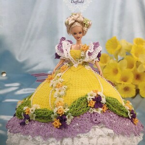 May include: A doll wearing a handmade yellow, white, and purple crochet gown with floral accents. The gown is part of the "Flower Garden Collection" and is labeled "Daffodil". The doll has blonde hair and is posed in front of a background with yellow flowers.