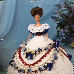 May include: A doll in a white crocheted gown from the "Flower Garden Collection." The dress features blue and white floral details, red trim, and bead embellishments. The doll has brown hair and is set against a blue background. The text "Forget-Me-Not" is visible.