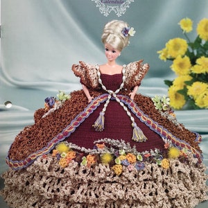 May include: A doll in a handmade crochet gown from the Flower Garden Collection. The gown has a burgundy bodice, brown and beige crochet skirt, and floral accents with tassels. The text "Annie's Glorious Gowns" is visible.