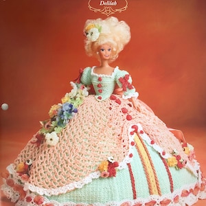 May include: A crochet doll wearing a green, pink, and white dress with a red and yellow trim. The doll has blonde hair and blue eyes. The dress is decorated with flowers and beads. The doll is part of the "Old South Collection" by Annie's Glorious Gowns.
