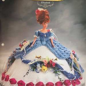 Può includere: Una bambola all'uncinetto che indossa un abito blu e bianco con un motivo floreale. La bambola indossa un nastro rosa intorno alla parte inferiore dell'abito. La bambola è intitolata "Elizabeth" e fa parte della "Collezione Old South di Annie's Glorious Gowns".