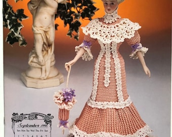 May include: A crocheted doll wearing a peach-colored dress with white lace trim and a matching hat with flowers. The doll is holding a purple and white crocheted umbrella. The doll is standing in front of a garden statue.