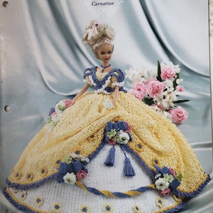 May include: A doll wearing a yellow and white crocheted gown with blue accents and floral embellishments. The gown is part of the "Flower Garden Collection" and is labeled "Carnation". The doll has blonde hair and is posed in front of a soft background.