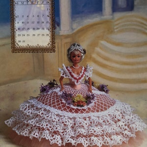 May include: A doll in a white and pink crocheted dress with a layered skirt. The doll wears a tiara and pearl necklace. A calendar with "Miss August 1998" is in the background. The doll is an Annie Potter Original.