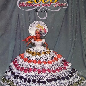 May include: A crochet doll wearing a white dress with multiple layers of colorful ribbons. The doll is wearing a white hat with a red ribbon and has blonde hair. The background is a sparkly gray fabric. The title of the pattern is "2003 Ribbons and Lace State Fair" and the designer is "Annie Potter Presents".