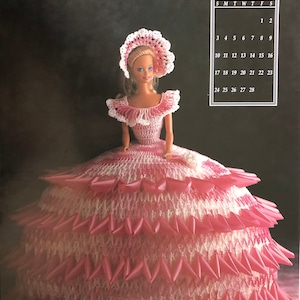 May include: A pink and white crocheted doll dress with a white crocheted hat. The doll is wearing a pink and white dress with a white crocheted hat. The dress has a layered skirt with pink ribbons. The doll is standing on a black background. The image also includes a calendar for February.