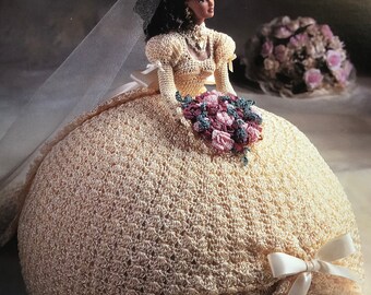 May include: A crocheted doll dressed in a white wedding gown with a veil and holding a bouquet of pink flowers. The doll is sitting on a white crocheted base with a white satin bow. The text "The Bridal Belle Collection" and "Miss June" is printed on the image.