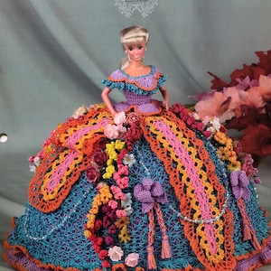 May include: A doll wearing a handmade crochet gown from the "Flower Garden Collection." The dress features a blue, orange, pink, and purple color scheme, adorned with floral embellishments and beaded accents. The text "Annie's Glorious Gowns" is at the top.