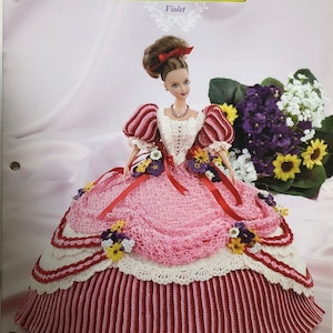 Puede incluir: Una muñeca de crochet con un vestido rosa y rojo con un borde de encaje blanco. La muñeca sostiene un ramo de flores moradas y amarillas. La muñeca se titula "Violeta" y forma parte de la "Colección Jardín de Flores".