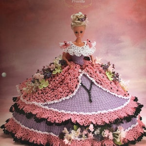 May include: A crochet doll wearing a pink and purple gown with a white lace collar and floral embellishments. The doll is titled "Priscilla" and is part of the "Annie's Glorious Gowns Old South Collection".