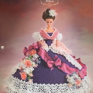 May include: A crochet doll dress pattern for a doll named Rebecca. The dress is purple with pink accents and white lace trim. The pattern is from Annie's Glorious Gowns Old South Collection.