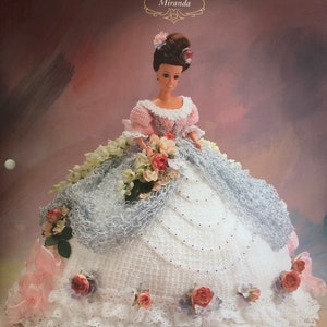 May include: A crocheted doll dress in white and pink with a lace overlay and floral embellishments. The doll is wearing a pink crocheted bodice and a white crocheted skirt. The dress is part of the "Annie's Glorious Gowns Old South Collection" and is named "Miranda".