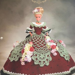 Könnte beinhalten: Eine Puppe trägt ein burgunderfarbenes Häkelkleid mit grüner Spitzenborte und floralen Akzenten. Das Kleid ist mit Perlen verziert und hat unten einen schwarzen und weißen Saum. Die Puppe hat blonde Haare und blaue Augen. Der Titel "Annie's Glorious Gowns Old South Collection Christina" ist oben im Bild gedruckt. Der Text "An Original Design by Annie Potter" ist unten im Bild gedruckt.