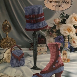 May include: Crocheted miniature riding boot, hat, and purse in shades of purple and burgundy. The boot has a high heel and a button detail. The hat has a wide brim and a ribbon detail. The purse has a flap closure. The items are displayed on a white background with a floral pattern. The text "Annie's Miniature Shoe Society" is visible in the upper right corner of the image.