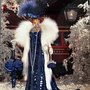 May include: A doll wearing a floor-length, dark blue sequined gown with a white faux fur stole, a matching blue hat with feathers, and a long silver necklace. The doll holds a blue parasol and a small blue handbag, labeled Paradise Publications Crochet Collector Costume Volume 100 1911 Orient Express Dinner Dress.
