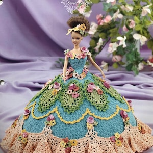 May include: A Barbie doll wearing a handmade crochet gown. The gown is blue with green, yellow, and pink floral accents. The doll's hair is styled in a bun with a floral headband. The text "Flower Garden Collection Sweet Pea" is visible.