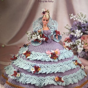 May include: A doll in a crocheted gown from the "Flower Garden Collection." The lavender dress has layers of light blue trim and is adorned with floral accents. The doll has blonde hair and is holding a bouquet.