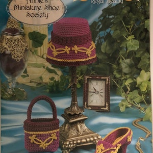May include: A crochet pattern for a purple and gold shoe, a matching handbag, and a hat. The pattern is titled "Annie's Miniature Shoe Society" and "Stepping Through Time Collection: Regal Beauty".