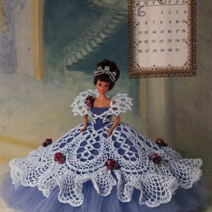 May include: A doll in a white crocheted dress with a blue bodice and a tulle skirt, decorated with red roses. The doll wears a tiara. A calendar for July 1998 is in the background. The dress is a work of art.