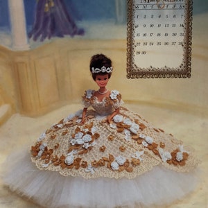 May include: A Barbie doll wearing a cream-colored gown with white and gold floral accents. The doll has a tiara and pearl necklace. A calendar for November 1998 is in the background.