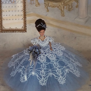 May include: A doll in a pale blue gown with white lace details and a bouquet of blue flowers. The doll has dark hair and is positioned in front of a painted backdrop with a calendar reading "1998 Miss February."