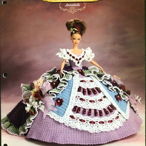 May include: A crochet doll dress pattern for the Old South Collection, featuring a doll wearing a purple, blue and white dress with a burgundy ribbon and floral accents. The pattern is called "Annabelle".