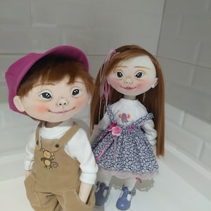 May include: Two handmade cloth dolls, one wearing a pink hat and brown overalls with a monkey design, the other wearing a blue and white floral dress with a pink flower and ribbon.