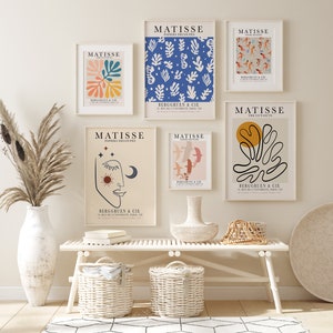 May include: Five framed art prints with a white background and black text. The prints feature colorful abstract designs and the text "MATISSE" and "PAPIERS D&Eacute;COUP&Eacute;S". The prints are hanging on a white wall above a white bench with two wicker baskets.