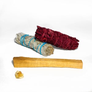 Puede incluir: Tres varitas de sahumerio, una blanca, una roja y una marrón, están dispuestas sobre una superficie blanca. La varita de sahumerio blanca está atada con una cuerda azul. La varita de sahumerio roja está atada con una cuerda roja. La varita de sahumerio marrón es un palo largo y delgado.