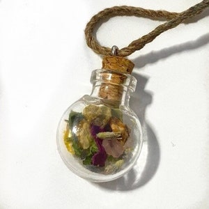 Op de afbeelding: Een ketting met een klein glazen flesje met een kurken stop. Het flesje is gevuld met gedroogde bloemen en kruiden, waaronder paars, roze, geel en groen.