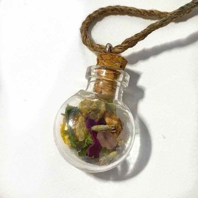Op de afbeelding: Een ketting met een klein glazen flesje met een kurken stop. Het flesje is gevuld met gedroogde bloemen en kruiden, waaronder paars, roze, geel en groen.