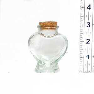May include: A clear glass heart-shaped bottle with a cork stopper. The bottle is approximately 3 inches tall.